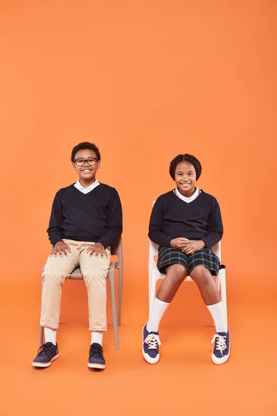 Felici scolari afro-americani in uniforme sorridenti e seduti su sedie su sfondo arancione — Foto stock