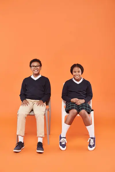 Happy african american schoolkids in uniform smiling and sitting on chairs on orange backdrop — Stock Photo