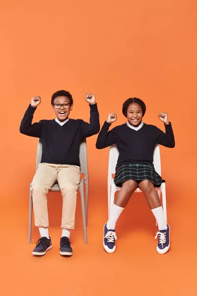 Animado Africano americano alunas em uniforme sorrindo e sentado em cadeiras em pano de fundo laranja — Fotografia de Stock