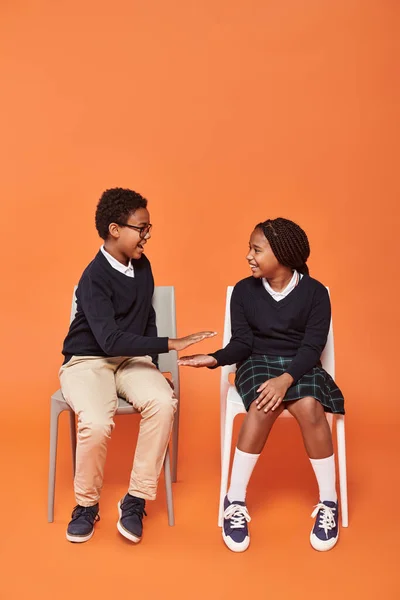 Alegre afroamericano colegiales en uniforme sentado en sillas y dando cinco en naranja telón de fondo - foto de stock