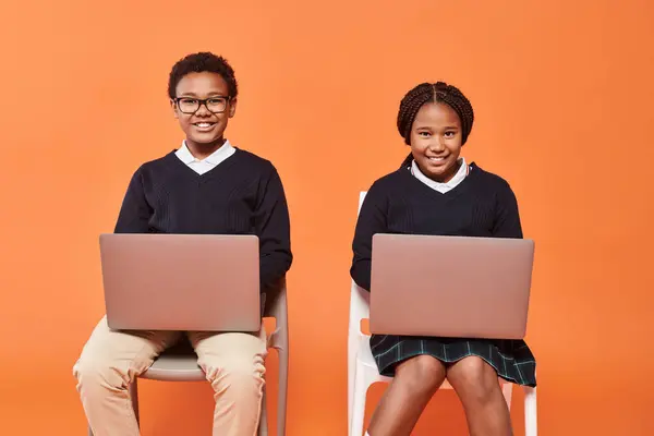 Happy african american schoolkids in uniform sitting on chairs and using laptops on orange backdrop — Stock Photo