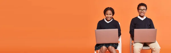 Happy African American schoolkids em uniforme sentado em cadeiras e usando laptops em laranja, banner — Fotografia de Stock