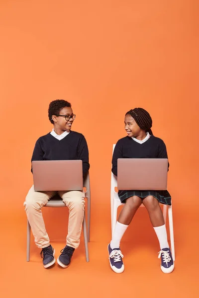 Niños afroamericanos felices en uniforme sentados en sillas y usando computadoras portátiles sobre fondo naranja - foto de stock