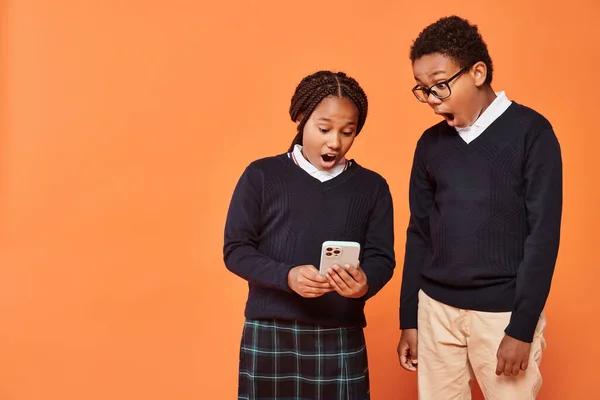 Emotional reaction, african american schoolkids in uniform looking at smartphone on orange — Stock Photo