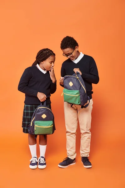 Réaction, écoliers afro-américains en uniforme tenant des sacs à dos et debout sur fond orange — Photo de stock