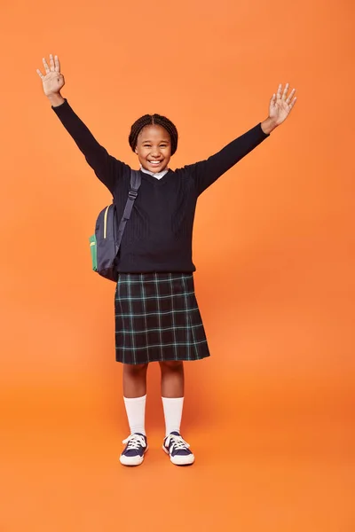 Cheerful african american schoolgirl in uniform smiling and holding backpack on orange background — Stock Photo