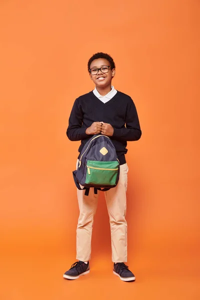 Happy african american schoolboy in uniform smiling and holding backpack on orange backdrop — Stock Photo