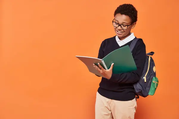 Happy african american schoolboy in uniform holding backpack and looking at textbook on orange — Stock Photo