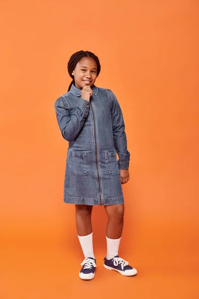 Повна довжина щасливої афроамериканської дівчини в джинсовій сукні позує і посміхається на помаранчевому тлі — стокове фото