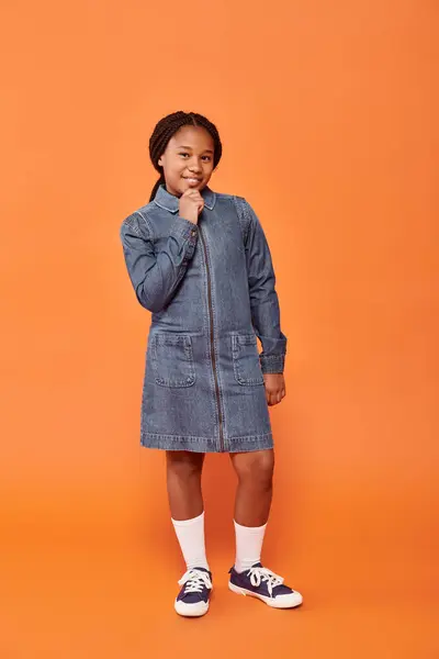 Full length of happy african american girl in denim dress posing and smiling on orange backdrop — Stock Photo