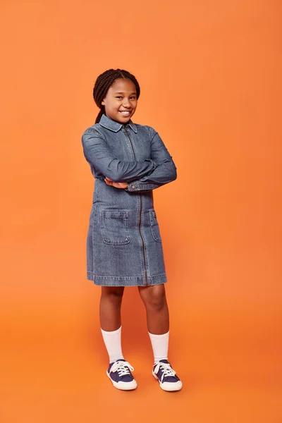 Full length of happy african american girl in denim dress posing with folded arms on orange backdrop — Stock Photo