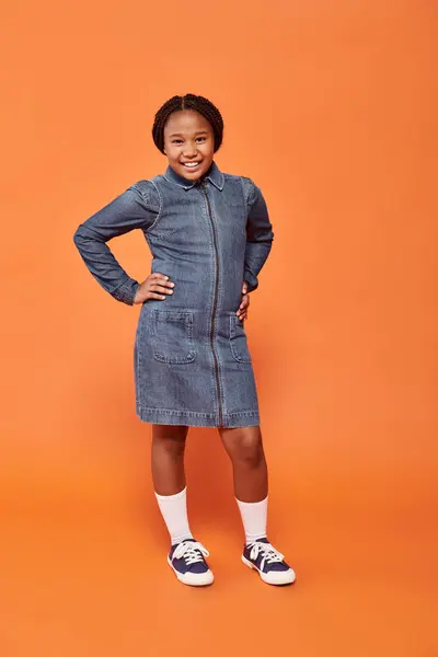 Pleine longueur de fille afro-américaine heureuse en robe de denim posant avec les mains sur les hanches sur l'orange — Photo de stock