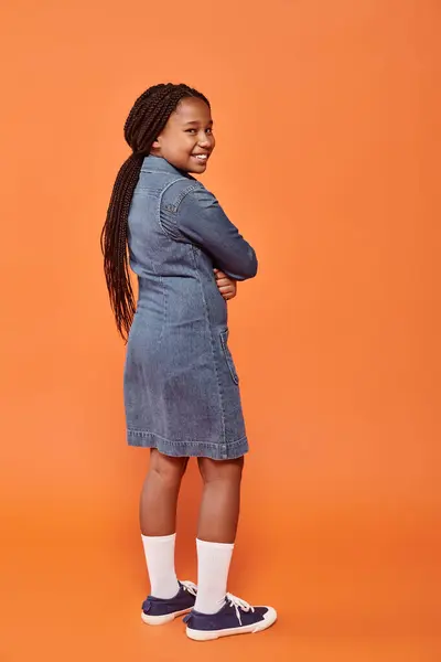 Comprimento total da menina americana africana positiva em vestido de ganga em pé sobre fundo laranja — Fotografia de Stock