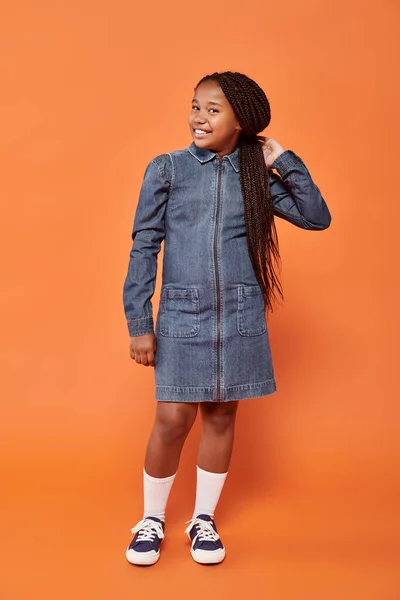 Comprimento total da menina americana africana positiva com tranças posando em vestido de ganga no fundo laranja — Fotografia de Stock