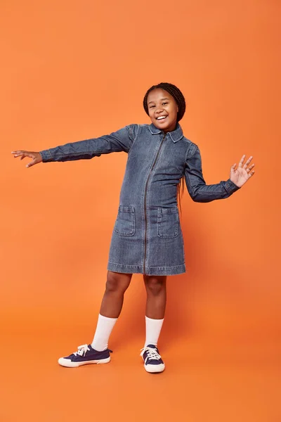 Pleine longueur de fille afro-américaine excitée avec des tresses posant en robe de denim sur fond orange — Photo de stock