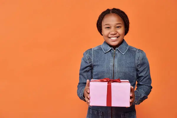 Happy african american girl in denim dress holding wrapped present and looking at camera on orange — Stock Photo