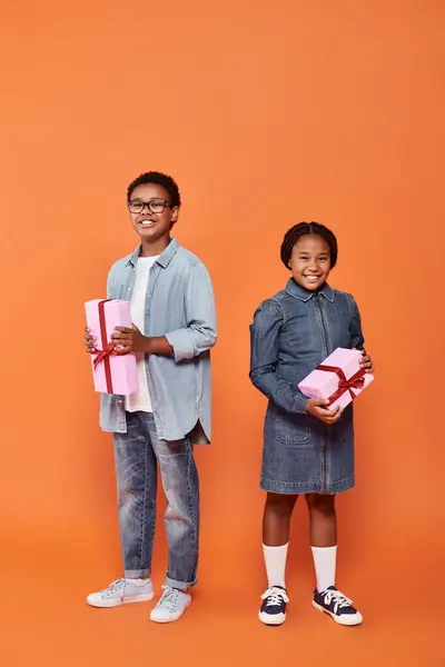 Niños afroamericanos felices sosteniendo regalos envueltos y de pie sobre fondo naranja - foto de stock