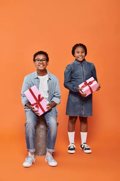 Niños afroamericanos alegres sosteniendo regalos envueltos y de pie sobre fondo naranja - foto de stock