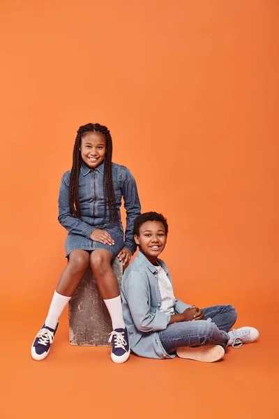 Crianças americanas africanas alegres em traje casual jeans sentados juntos no fundo laranja — Fotografia de Stock