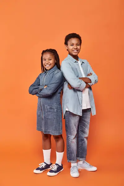 Happy african american children in casual denim attire standing with folded arms on orange backdrop — Stock Photo