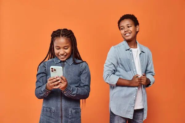 Glücklich afrikanisch-amerikanische Mädchen mit Zöpfen im Jeanskleid mit Smartphone in der Nähe von Jungen auf orangefarbenem Hintergrund — Stockfoto