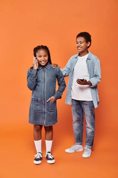 Happy african american girl and boy in casual wear using smartphones on orange background — Stock Photo