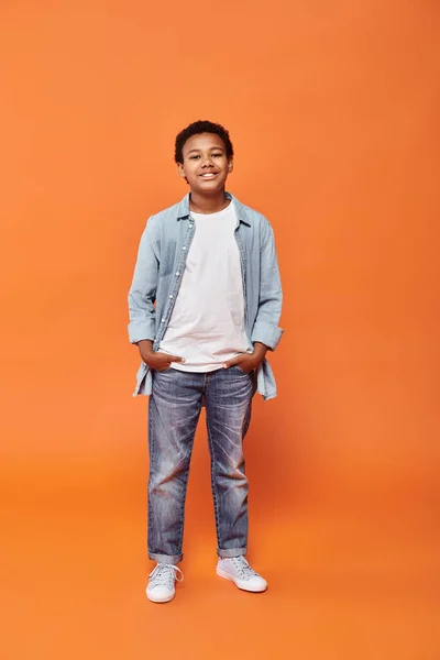 Cheerful preteen african american boy in casual outfit with hands in pockets on orange backdrop — Stock Photo