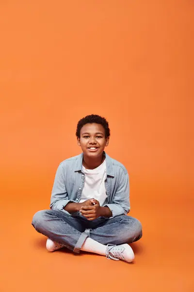 Gai préadolescent afro-américain garçon en tenue décontractée assis sur le sol avec les jambes croisées — Photo de stock