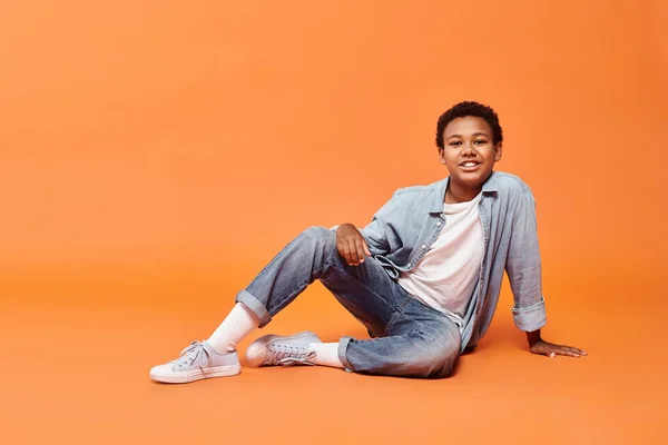 Happy african american boy in everyday wear posing on floor on orange backdrop and smiling at camera — Stock Photo