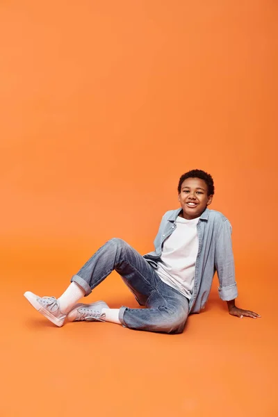 Alegre preadolescente afroamericano chico sentado en el suelo y sonriendo felizmente a la cámara - foto de stock