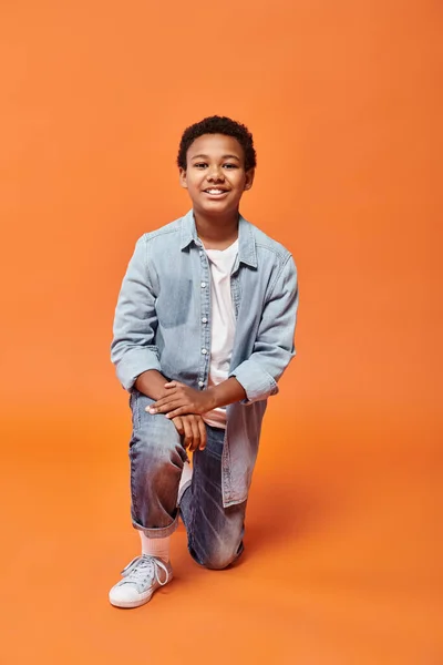 Веселий афроамериканський хлопчик у повсякденному вбранні, що стоїть на одному коліні і щасливо посміхається на камеру — стокове фото