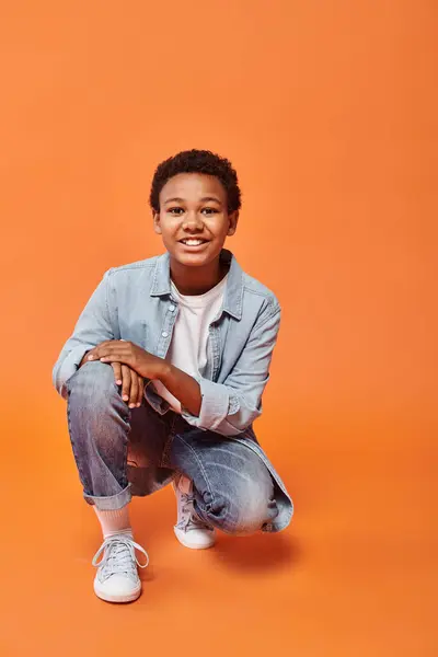 Cheerful preteen african american boy in casual outfit standing on one jnee and smiling at camera — Stock Photo