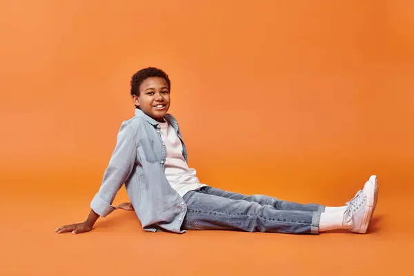 Joyeux preteen arican américain garçon en tenue quotidienne assis sur le sol et souriant à la caméra — Photo de stock
