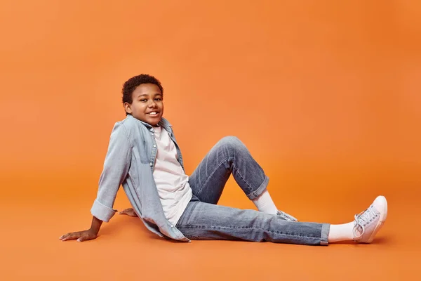 Happy preadolescent african american boy in casual attira sitting on floor on orange background — Stock Photo