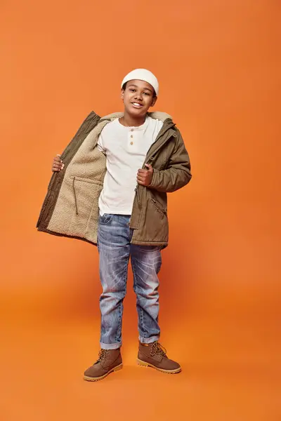Happy african american boy in winter warm jacket posing on orange backdrop and smiling at camera — Stock Photo