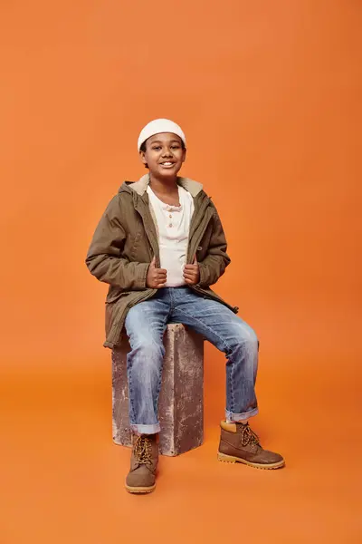 Alegre preadolescente afroamericano chico en invierno traje sentado y sonriendo alegremente a la cámara - foto de stock