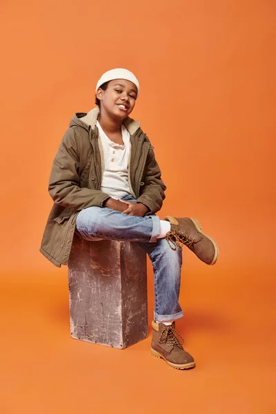Happy african american boy in winter jacket and jeans sitting and smiling joyfully at camera — Stock Photo