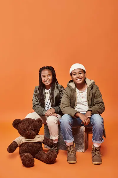 Alegres niños afroamericanos en trajes cálidos posando con oso de peluche sobre fondo naranja - foto de stock