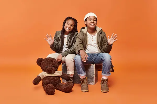 Jolly preteen bambini africani americani in abito invernale in posa con orsacchiotto e agitando alla macchina fotografica — Foto stock