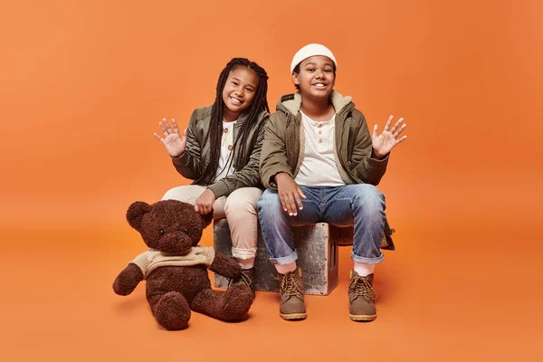 Jolly preteen african american children in winter outfit posing with teddy bear and waving at camera — Stock Photo
