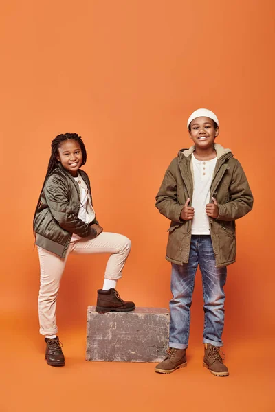 Joyous african american children in winter outfits posing on ornage backdrop and smiling happily — Stock Photo