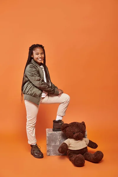 Cheerful preteen african american girl with braids in winter attire posing next to her teddy bear — Stock Photo