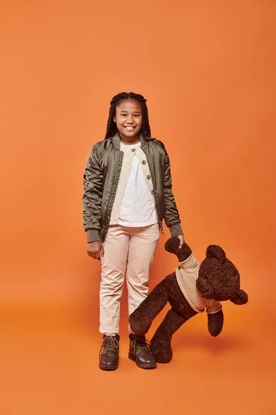 Alegre afroamericana chica con trenzas posando felizmente con su osito de peluche sobre fondo naranja - foto de stock