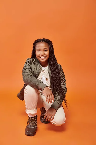Allegra ragazza afroamericana con trecce in giacca invernale accovacciata e sorridente alla macchina fotografica — Foto stock