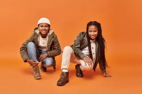 Cheerful preteen african american boy and girl in winter jackets squatting on orange backdrop — Stock Photo
