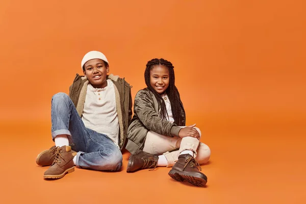 Happy preteen african american boy and girl in warm outfits sitting on floor and smiling at camera — Stock Photo