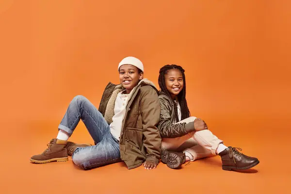 Alegre preadolescente afroamericano niños en invierno trajes sentado en el suelo sonriendo a la cámara - foto de stock