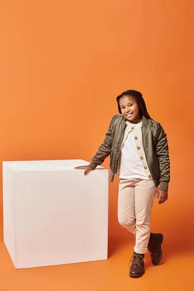 Alegre africano americano chica en invierno chaqueta posando cerca blanco cubo y sonriendo a cámara - foto de stock