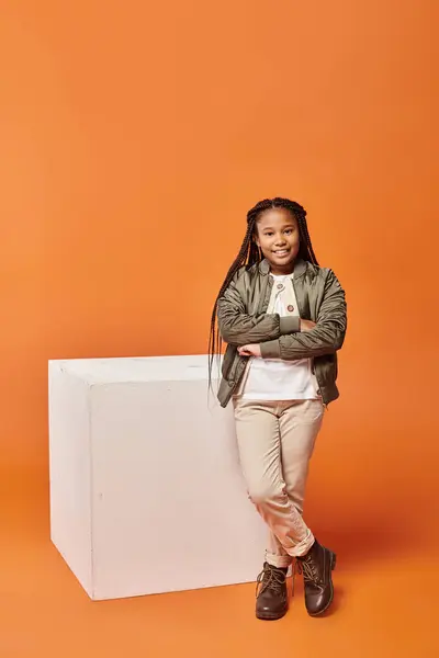 Joyeux afro-américaine fille en tenue chaude posant à côté de cube blanc avec les bras croisés sur la poitrine — Photo de stock