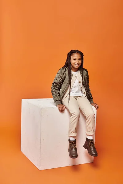 Happy african american girl in winter attire smiling at camera on white cube on orange backdrop — Stock Photo